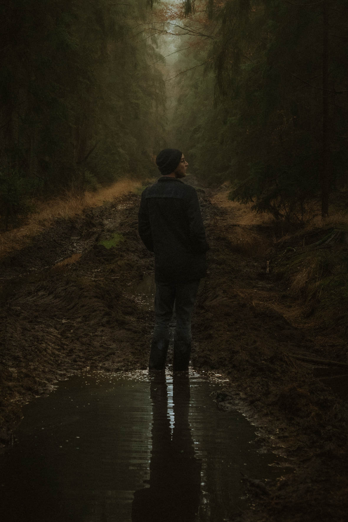 Dark autumn forest with a man standing in a puddle of a muddy forest road